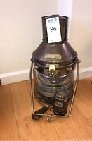 Full view of vintage Anchor copper and brass nautical oil lantern with glass burner section and attached small brass oil container and black rubber priming bulb pump on wooden floor.