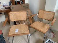 Two vintage cantilever arm chairs with woven rattan seats and backs, wooden frames and chrome bases placed together on carpeted floor.