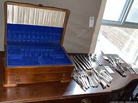 Open wooden two-level flatware box with blue velvet and cream satin lining alongside scattered flatware pieces