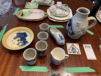 Overview of all 9 ceramic pieces including bowl, jug, 4 shot glasses, clog, creamer, and KLM tile on table.