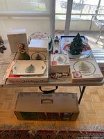 Full view of table with Spode Christmas Tree dinnerware boxes, ceramic tree light, snowman figurine, and boxed faux Christmas tree beneath table.