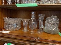 Shelf containing crystal and glass bowls, vase, decanter, and utensil rests arranged side by side showing detailed cut crystal design and glass transparency.