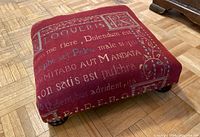 Angled side view showing red fabric upholstery with white and blue text motif and dark wooden bun feet with brass nailheads.