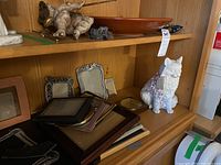 Shelf showing multiple assorted picture frames in wood and metal, ceramic cat figure and other small objects