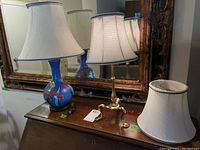 Two table lamps and one extra shade on a wooden table with mirror in background. Taller lamp has blue floral ceramic base and fabric shade. Smaller lamp has gold-toned metal base and fabric shade matching extra shade.