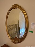 Oval gold framed beveled mirror hung on wall, showing top floral decoration and partial room reflection