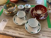 Overview photo showing all six items on a surface with green tape markings: red/gold tea cup and saucer, green wreath tea cup and saucer, two floral egg coddlers, and two white green leaf espresso cups and saucers.