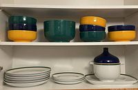 Two-shelf display showing multicolor stacking bowls, large green bowl on top shelf; dinner and side plates, covered soup tureen on platter below
