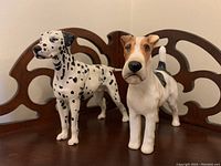 Front view of both dog figurines showing Dalmatian and Fox Terrier standing poses on wooden surface with decorative wooden back panel.