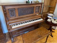 Front side view of Smith and Barnes upright piano with carved applique panels on the top front with floral designs, oak wood finish, and piano bench in front.