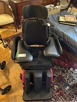 Front view of black Power Plus Mobility wheelchair showing padded neck, arm, and leg supports with leather-like cushioning and contoured headrest.