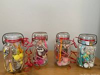 Four quart glass jars filled with various small toys and prizes, each with red ribbons around the necks, including a metallic handle on one lid and several colorful small items visible through the glass.