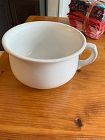 White ceramic antique chamber pot showing entire bowl and handle, placed on wooden surface with folded cloth in background.