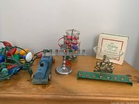 Photo of holiday items including two menorahs, a jar filled with vintage bulbs, a string of vintage bulbs, a wooden toy train engine, and Avon compote with box arranged on a wooden surface.