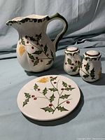 Large ceramic pitcher, round trivet, and salt and pepper shakers with holly berry design together on a cloth background