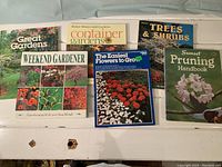 Photo of six softcover gardening books arranged on a white table. Titles visible include Great Gardens Weekend Gardener, Container Gardens by Better Homes and Gardens, Trees & Shrubs, Sunset Pruning Handbook, and The Easiest Flowers to Grow.