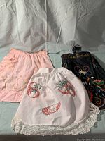 Front view of three half aprons laid flat showing white, pink, and black aprons with embroidery and lace details.
