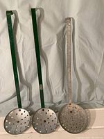 Three metal strainers with long handles standing upright against a white backdrop. Two have green painted handles, one with a bare metal handle. Each strainer has a circular perforated bowl.