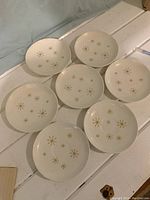 Top-down group photo showing 7 'Atomic Star' dishes with gold starburst patterns, 5 plates and 2 saucers with white ceramic finish.
