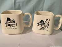 Photo of two cream-colored square ceramic mugs with black comic illustrations and captions by Herb Gardner's Nebbishes, placed side by side on white surface with light blue backdrop.
