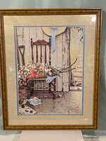 Full view of the framed lithograph artwork showing a wooden chair with a basket of colorful flowers and gardening items on a porch.