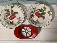 Two ceramic plates with painted red apples and white reticulated edges, placed beside a red apple cutter with stainless steel blades.