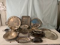 Photo displaying a variety of silverplate trays, bowls, and serving dishes arranged on a draped table, showcasing different shapes and sizes including round, oval, and rectangular trays with ornate detailing.