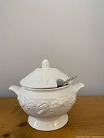 White ceramic Sonoma soup tureen with lid featuring embossed grapevine design and grape cluster handle. Includes metal ladle resting in tureen opening.