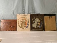 Four vintage photo albums arranged in a row showing front covers: one wooden textured cover with illustration and title, two with decorative winter scene cutouts on paper covers (cream and dark brown), and one corkboard-style cover.