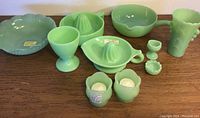 Full set of jadeite green glass items arranged on wood surface showing two juicers, a handled pot with spout, eye wash cup, salt bowl, two votive holders, a small vase, a drinking glass, and a rounded bowl with scalloped edge.