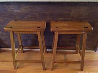 Front view of two wooden counter stools side by side, showing rectangular tops with dark joint insets and straight legs with footrest bars.