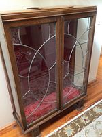 Front view of wooden display cabinet with leaded glass doors featuring inset silver metal geometric design and velvet red interior lining.