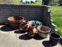Wide shot of all items: two wooden garden buckets, two terracotta strawberry planters, galvanized tub, black plastic planter, and watering can arranged against brick wall in sunlight.