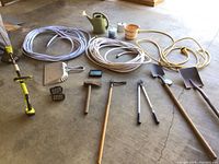 Overview of garden supplies including hoses, shovels, clippers, small axe, a watering can, and more arranged on a concrete floor.