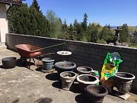 Wide view of the entire gardening lot including wheelbarrow, pots, bird bath, rake, hose barrel, and soil bags.