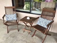 Two weathered teak folding chairs with a small matching teak folding table between them, all placed on a concrete floor outdoors