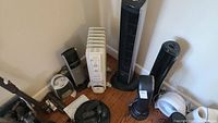 Photo showing multiple fans, heaters, and air filter units arranged on the floor including a white radiator heater and two black tower fans