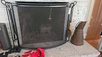 Wide shot of black metal fireplace screen with curved design, adjacent copper bucket, and small broom hanging beside screen