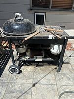 Weber grill on wheels with black domed kettle lid and prep table, showing grilling utensils atop and propane tank attached.
