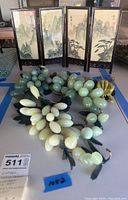 Photo showing three stone grapes clusters with varying green and white tones, leaves, and small yellow flower in front of a miniature folding silk art screen with four mountain landscape panels.