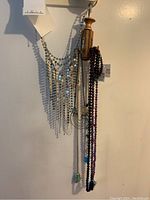 Three necklaces hanging on a wooden piece, one with multiple strands of silver metal discs, another with purple beads and green stone pendants.