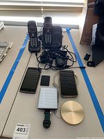 Full lot view showing two VTech cordless phones with base, two smartphones, silver external battery charger, white attachable flashlight, and small accessory on a white surface.