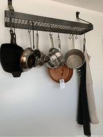 Kitchen pans hanging from a wall-mounted metal rack including cast iron grill pan, stainless steel pans, a copper pan, and two aprons on side.