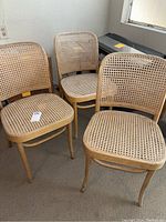 Three wicker Prague chairs showing woven rattan seats and backs, light wooden frames in a standing group, on carpeted floor near a wall.