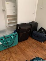Photo showing 4 travel bags lined up on floor: teal travel tote, black soft-sided suitcase, black hard-shell Samsonite suitcase, and dark blue carry-on bag. Clear plastic storage bin partially visible on left.