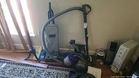 Photo of Royal upright vacuum with beige cloth bag and long hose standing beside a wall with other vacuums and accessories nearby.