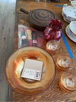 View of vintage marigold glass bowl, three matching cups, antique cast iron bed warmer, cranberry glass pitcher, and bag of chopsticks on a wooden table.