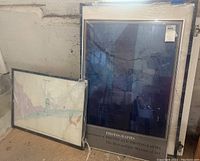 Two framed art pieces leaning against a wall; one is a large photographic print with a blue abstract architectural design from the Metropolitan Museum of Art; the other is a smaller framed image wrapped in bubble wrap with partially visible white matte.