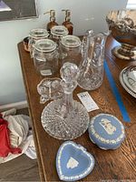 Photo showing Wedgwood blue porcelain trinket dishes, cut crystal decanter with stopper, cut crystal pitcher, glass jars with clamp lids, amber glass pump bottles, and small divided dish arranged on wooden surface.
