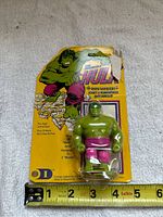 Front view showing the green mini Hulk figure in purple shorts inside the blister pack attached to yellow cardboard backing. Tape measure at base shows size.